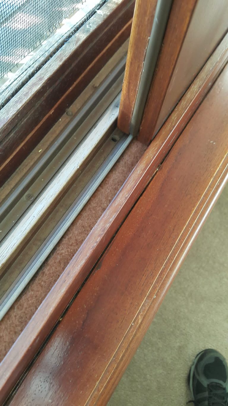 Ventana de madera barnizada abierta con mosquitero negro exterior. La ventana corrediza muestra aislamiento con burletes de fieltro naranja claro en la junta inferior y el espacio entre el riel y el marco.