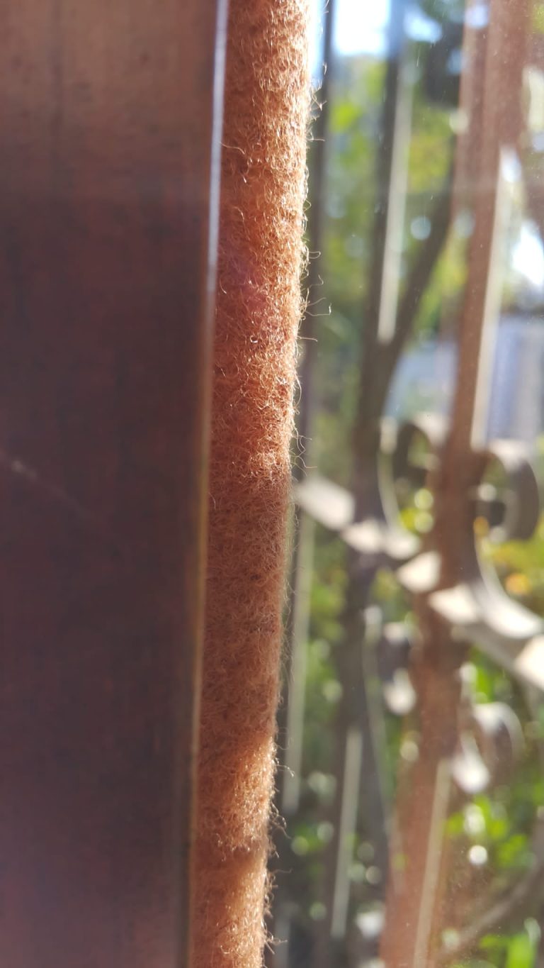 Primer plano de un burlete de fieltro marrón claro, instalado en el marco lateral de una abertura, proporcionando aislamiento eficaz.